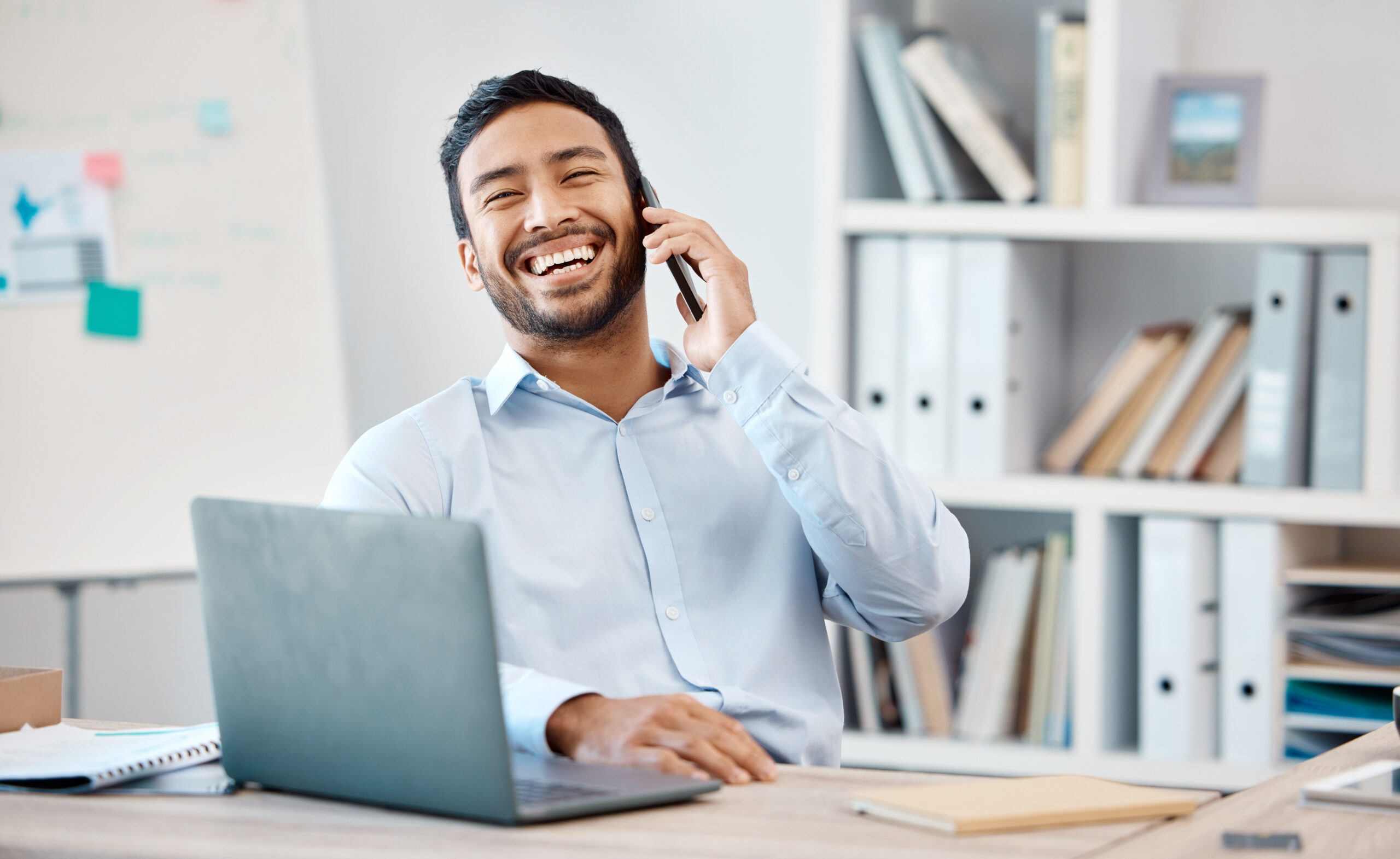 Happy businessman on a phone call while working on a laptop at the desk in his modern office. Corporate, professional and company manager laughing while having a mobile conversation with technology