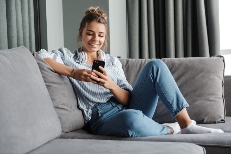 Junge Frau im gestreiften Hemd lächelt und liest auf ihrem Smartphone über versteckte Geschäftskosten, während sie sich auf einem grauen Sofa entspannt.