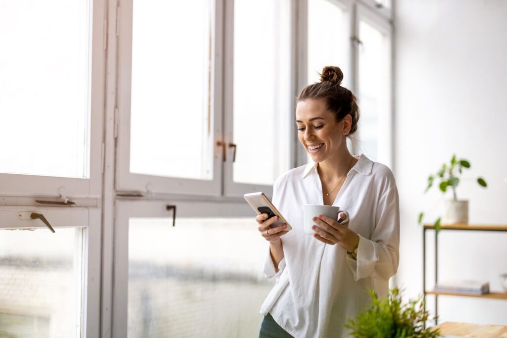 Frau lächelt in ein Smartphone an einem großen Fenster und hält eine Tasse in der Hand; lässiger, heller Raum mit Pflanzen - möglicherweise liest sie über versteckte Geschäftskosten.