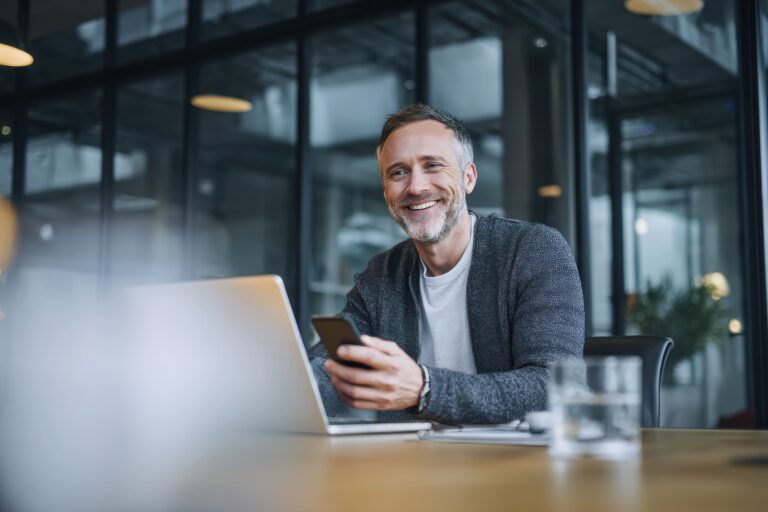 Bärtiger Mann, lächelnd am Schreibtisch mit Laptop, Smartphone in der Hand, in einem modernen Büro mit Glaswänden - möglicherweise prüft er versteckte Geschäftskosten.