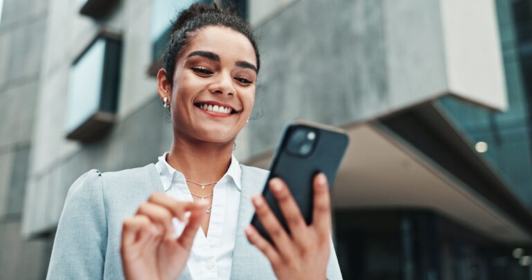 Eine lächelnde Frau in einem grauen Blazer benutzt ihr Smartphone vor einem modernen Gebäude und blickt selbstbewusst auf den Bildschirm, der durch eine gute Netzabdeckung ermöglicht wird.