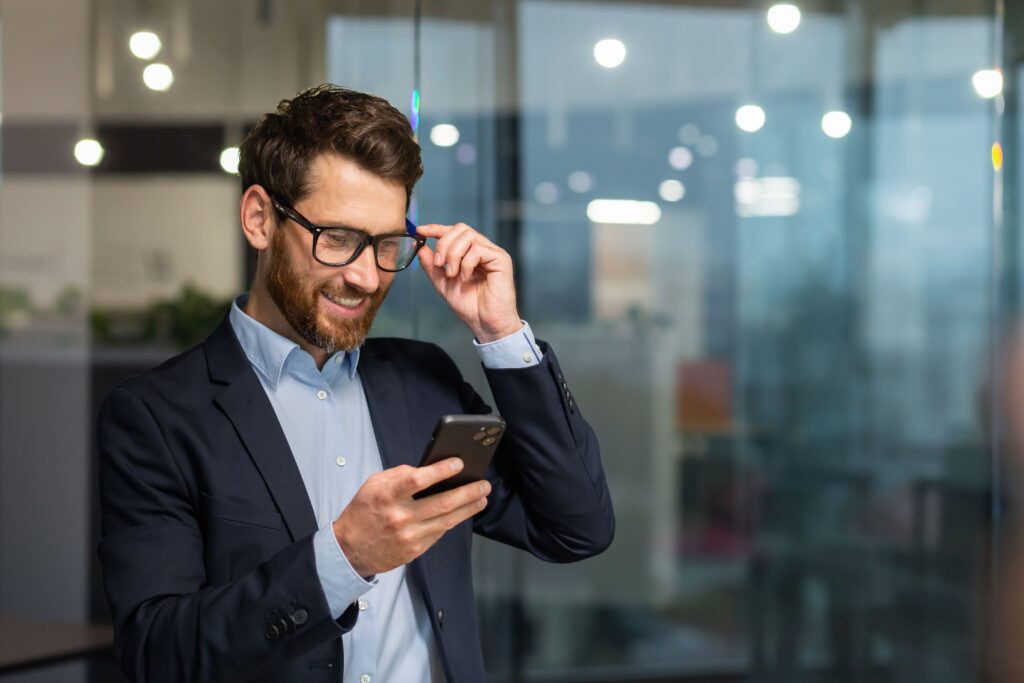 Ein lächelnder Mann im Anzug und mit Brille überprüft sein Smartphone in einem modernen Glasbüro, verschwommene Lichter suggerieren Cloud Computing.