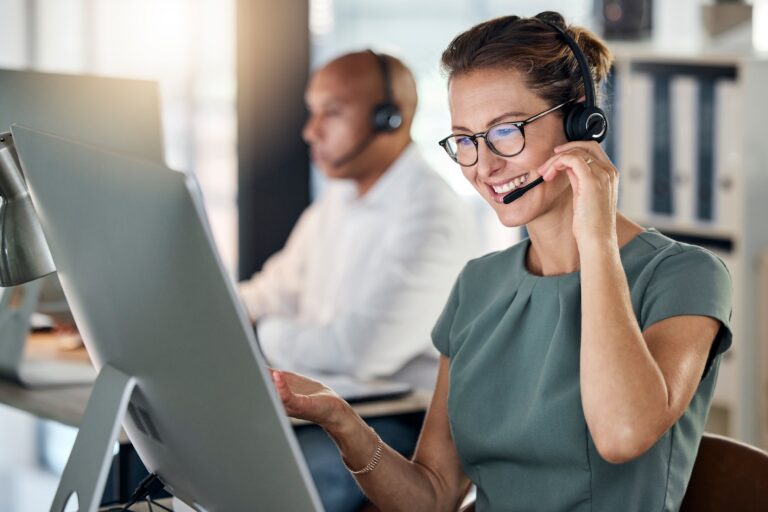 Frau mit Brille und Headset arbeitet lächelnd im Büro am PC, Kollege mit Headset im Hintergrund.