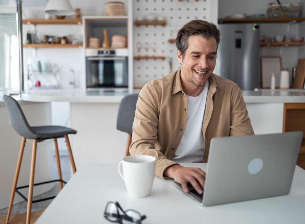 Lächelnder Mann am Küchentisch benutzt Laptop mit Becher und Brille, genießt flexible Geschäftstarife in einer modernen Küche.