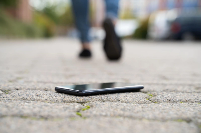 Smartphone mit dem Gesicht nach oben auf dem Bürgersteig; im Hintergrund geht eine unscharfe Person weg.