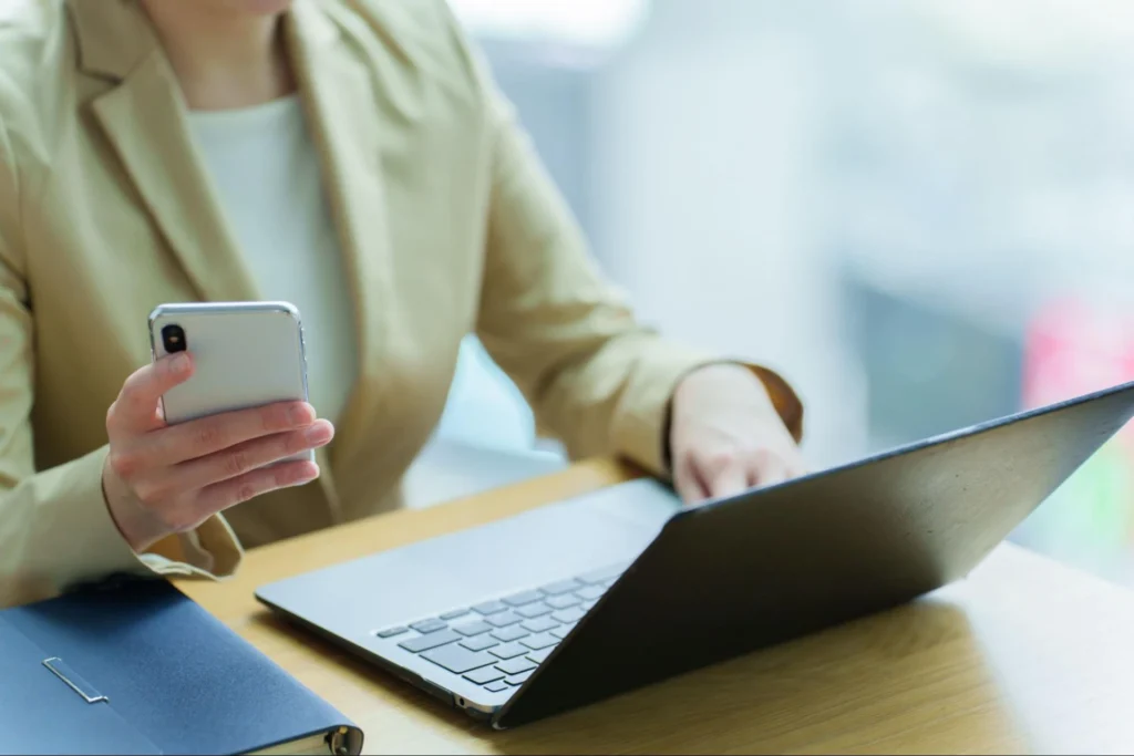 Person in beigem Blazer prüft Smartphone und arbeitet am Laptop am Schreibtisch; geschlossenes Notebook liegt daneben.