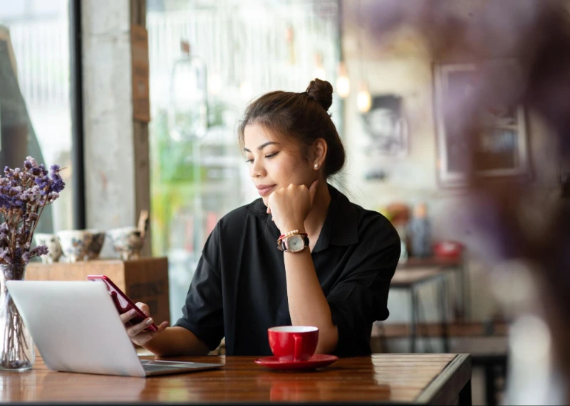 Frau am Kaffeetisch, lächelnd auf ihr Telefon. Laptop und rote Kaffeetasse im Vordergrund, unscharfe Blumen im Hintergrund.