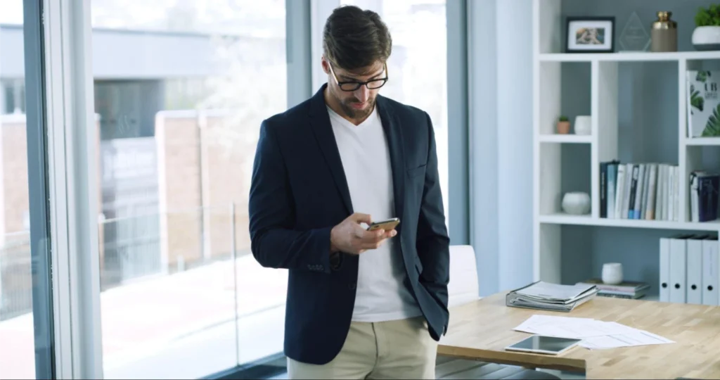 Mann mit Brille und Geschäftskleidung steht in einem Büro und schaut auf sein Smartphone; hinter ihm Regale, Schreibtisch, Papiere und Fenster.