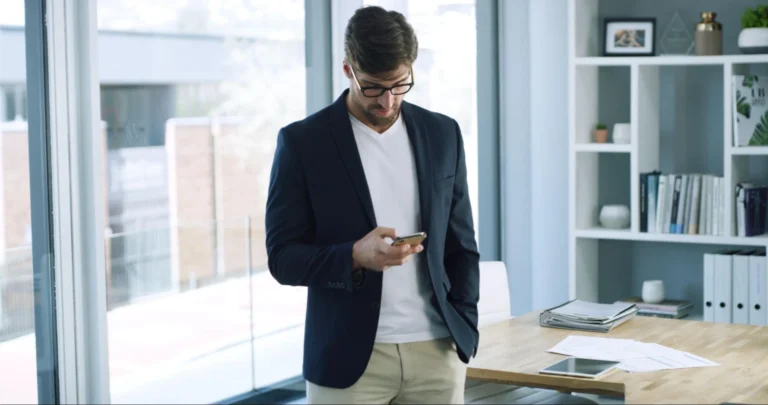 Mann mit Brille und Geschäftskleidung steht in einem Büro und schaut auf sein Smartphone; hinter ihm Regale, Schreibtisch, Papiere und Fenster.