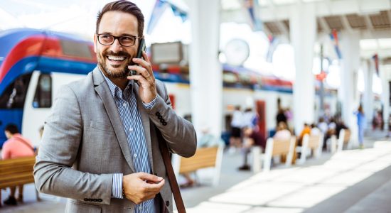 Businessman using mobile phone at train station. Handsome young man on business trip walking with his luggage and talking on cellphone at train station. Travelling businessman making phone call